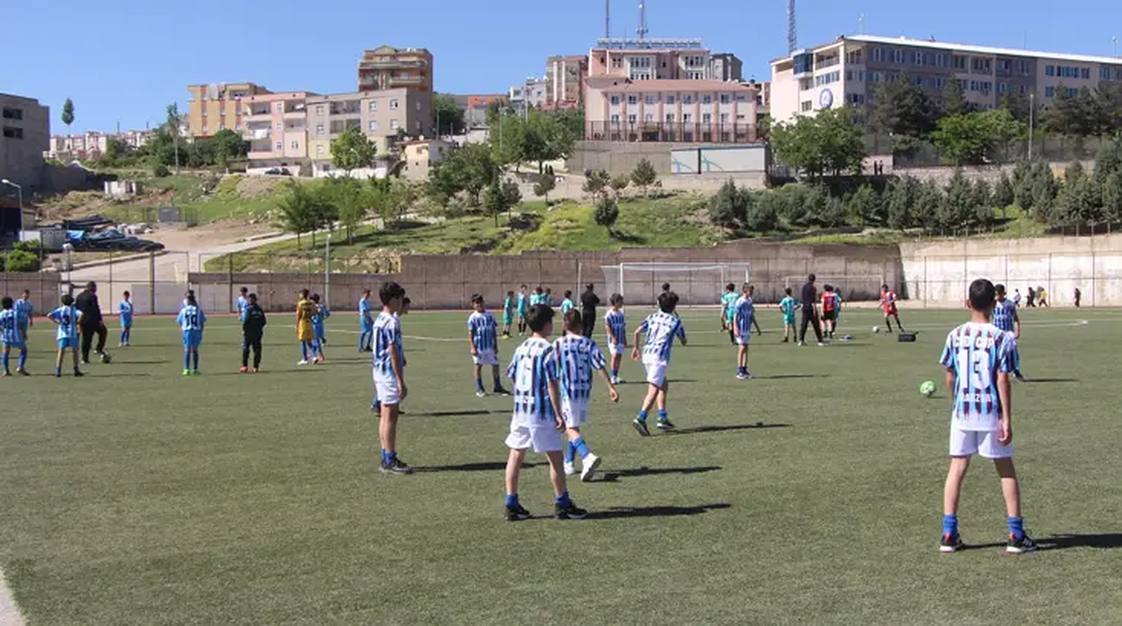 Gün ışığında bir futbol sahasında mavi beyaz çizgili formalı çocuklar; arkada yamaçta binalar